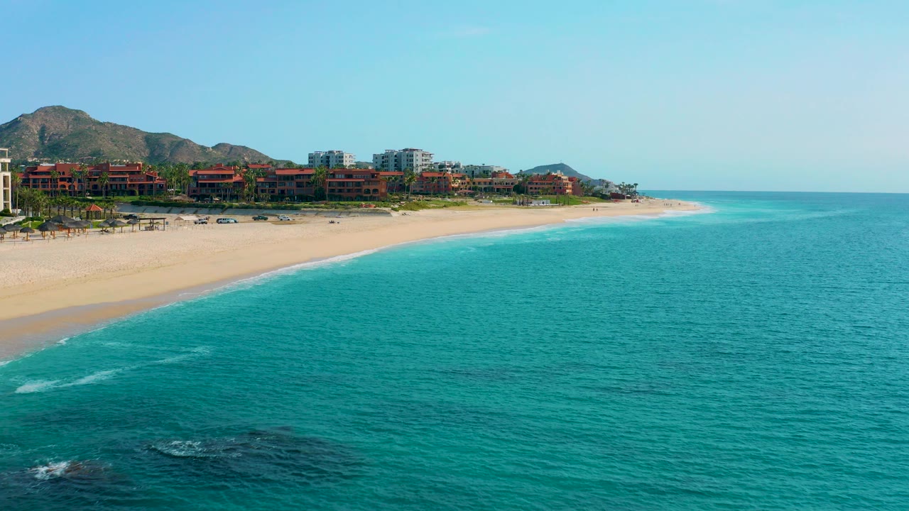 Opening aerial frame over Desert Park Natural Reserve in Baja California Sur, Mexico, showing turquoise water, a pale sand beach, resort buildings, mountain foothills, and no visible people.