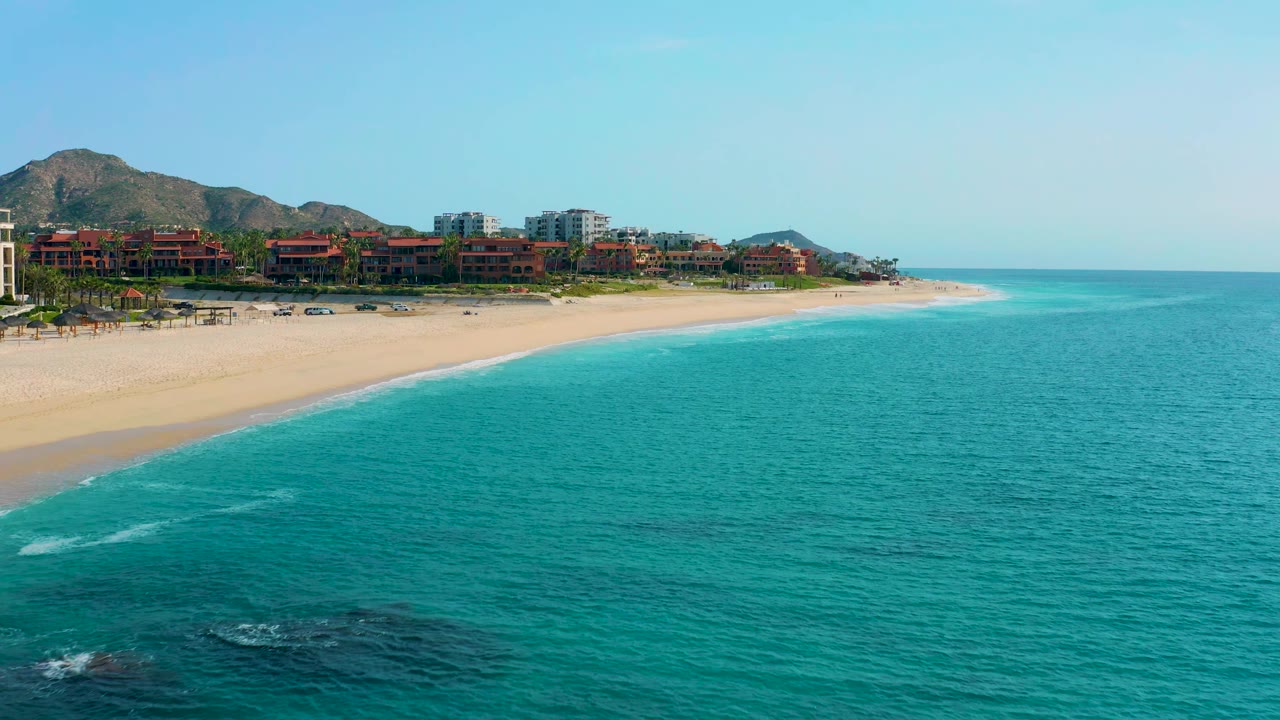Second frame holds the Desert Park Natural Reserve coastline wider across the center, with rippled turquoise water, a curving sand beach, resort buildings, mountain foothills, and no visible people.