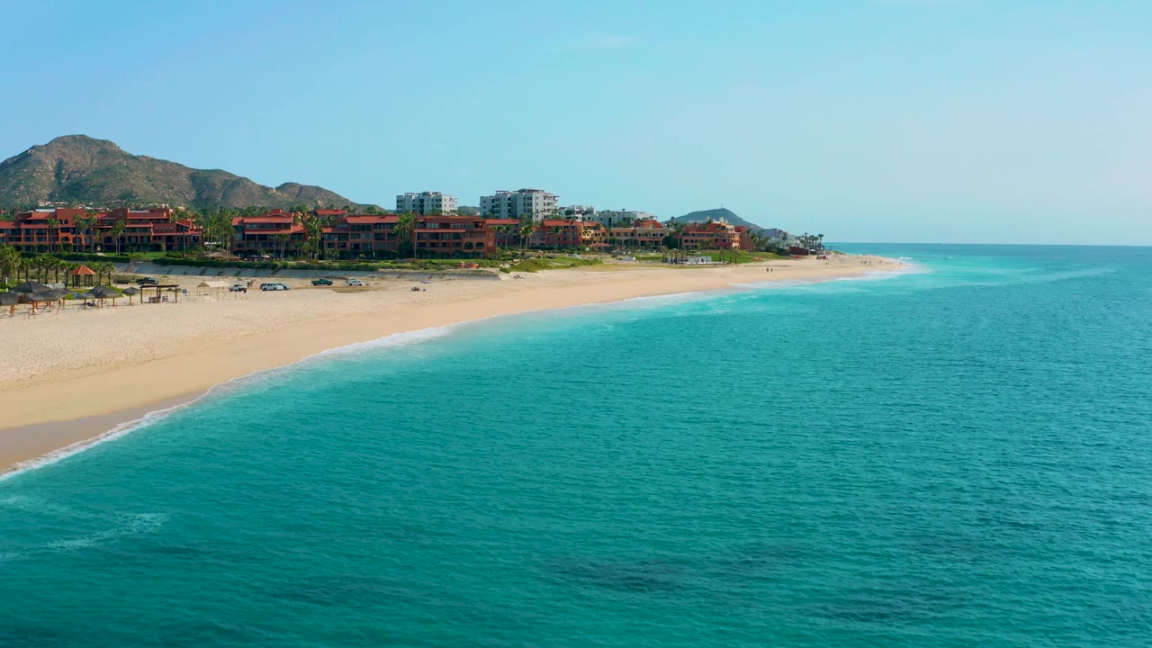 Turquoise ocean and sandy beach with resort buildings below brown mountains.