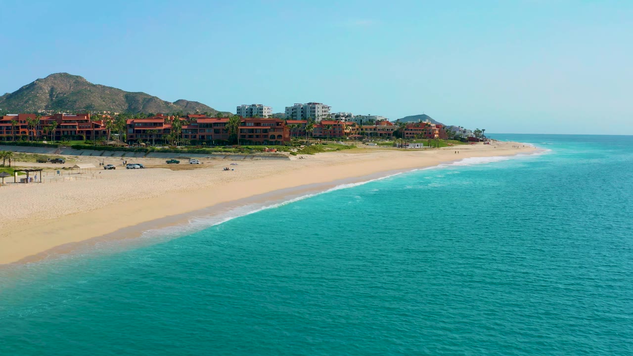 Aerial frame in Desert Park Natural Reserve, Baja California Sur, showing the curved sandy shoreline, teal ocean, resort buildings, mountains, and no visible people.