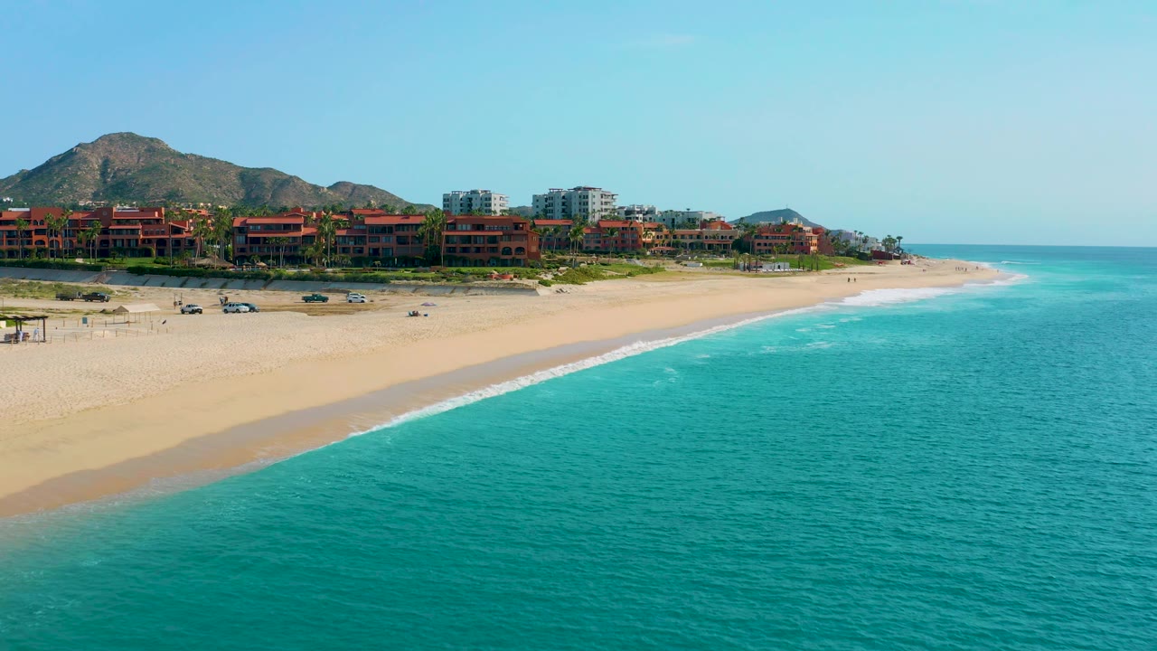 Wide coastal aerial in Desert Park Natural Reserve, Baja California Sur, with turquoise water dominating the foreground, a pale sand beach, resort blocks, mountains, and no visible people.