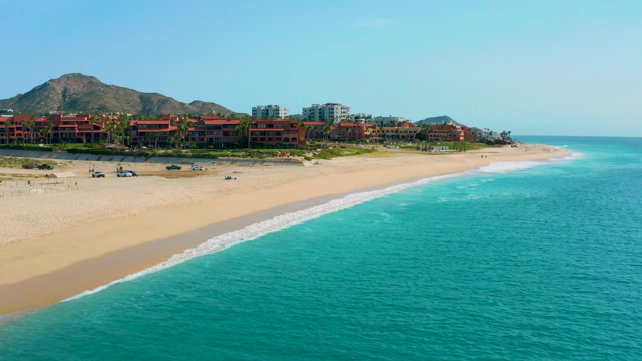 High-angle beach view in Desert Park Natural Reserve, Baja California Sur, with white surf reaching the sand, resort buildings along the upper beach, mountains, and no visible people.