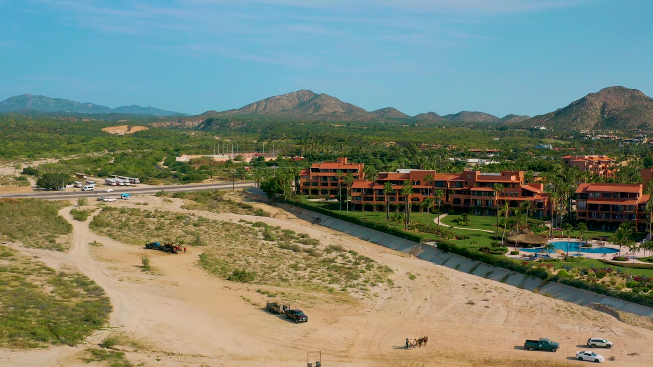 Wide aerial over Desert Park Natural Reserve in Baja California Sur showing sandy desert tracks, terracotta resort buildings, a blue pool, green palms, distant mountains, and no visible people.