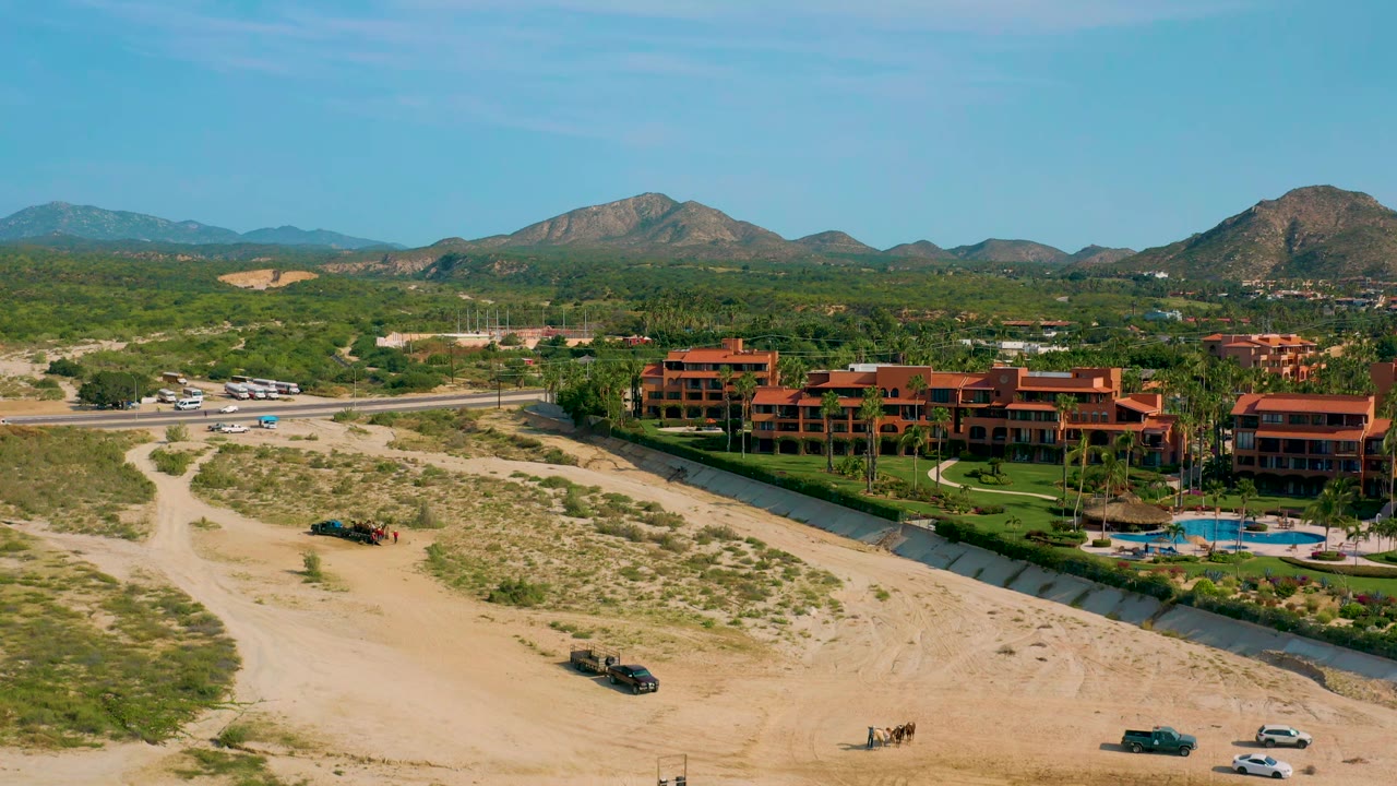 Aerial frame in Baja California Sur with the resort shifted toward center, dirt paths and scrub in front, a road with parked vehicles beyond, mountains on the horizon, and no visible people.