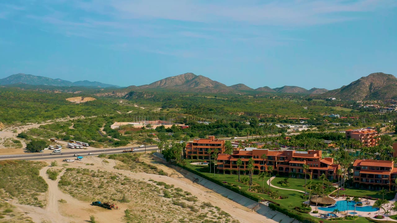 Aerial frame in Baja California Sur with the resort shifted slightly lower in view, showing red-orange buildings, landscaped lawns, pool water, a road with vehicles, scrubland, mountains, and no visible people.