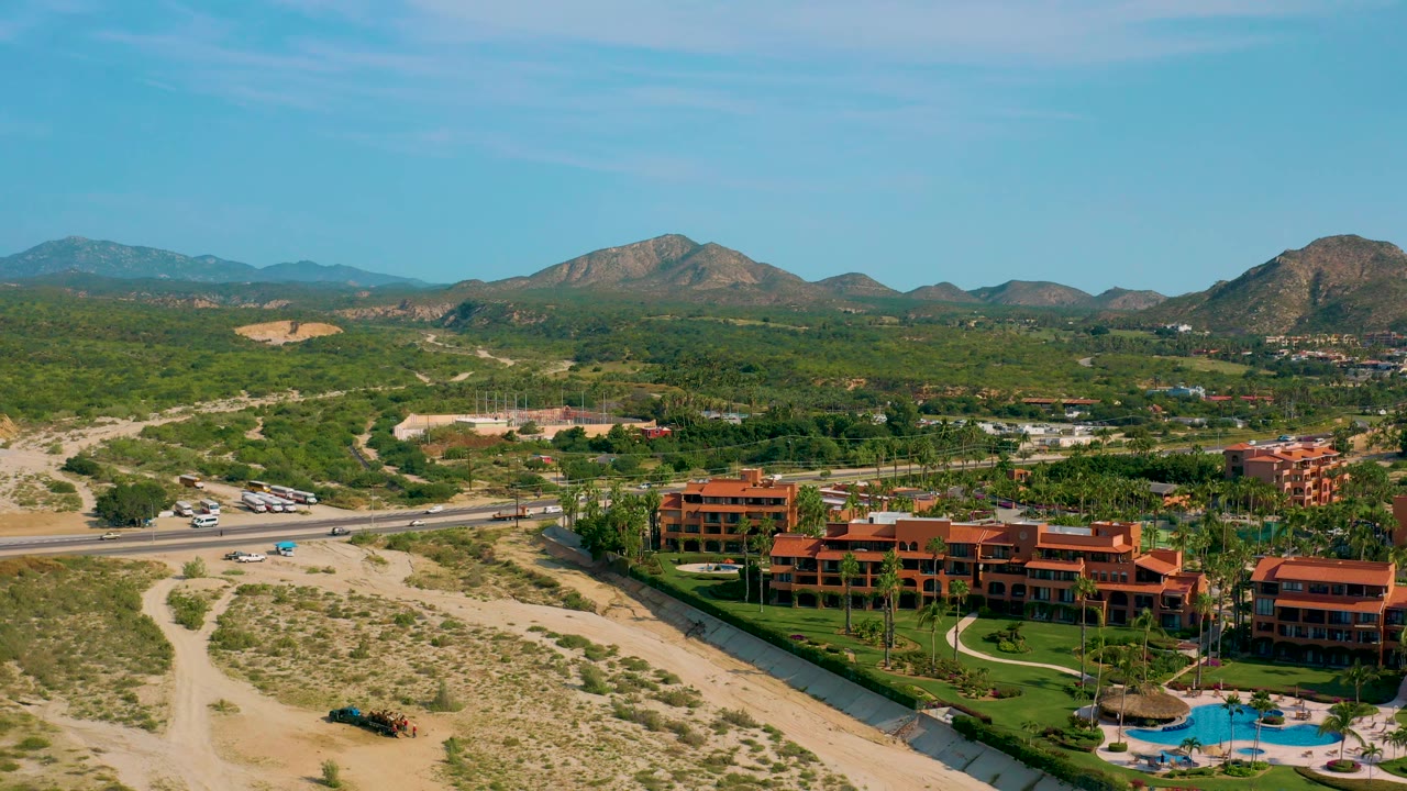 High-angle view in Desert Park Natural Reserve, Baja California Sur, Mexico, with the concrete canal cutting past the resort, vehicles on the highway, sandy open ground, green hills, distant mountains, and no visible people.