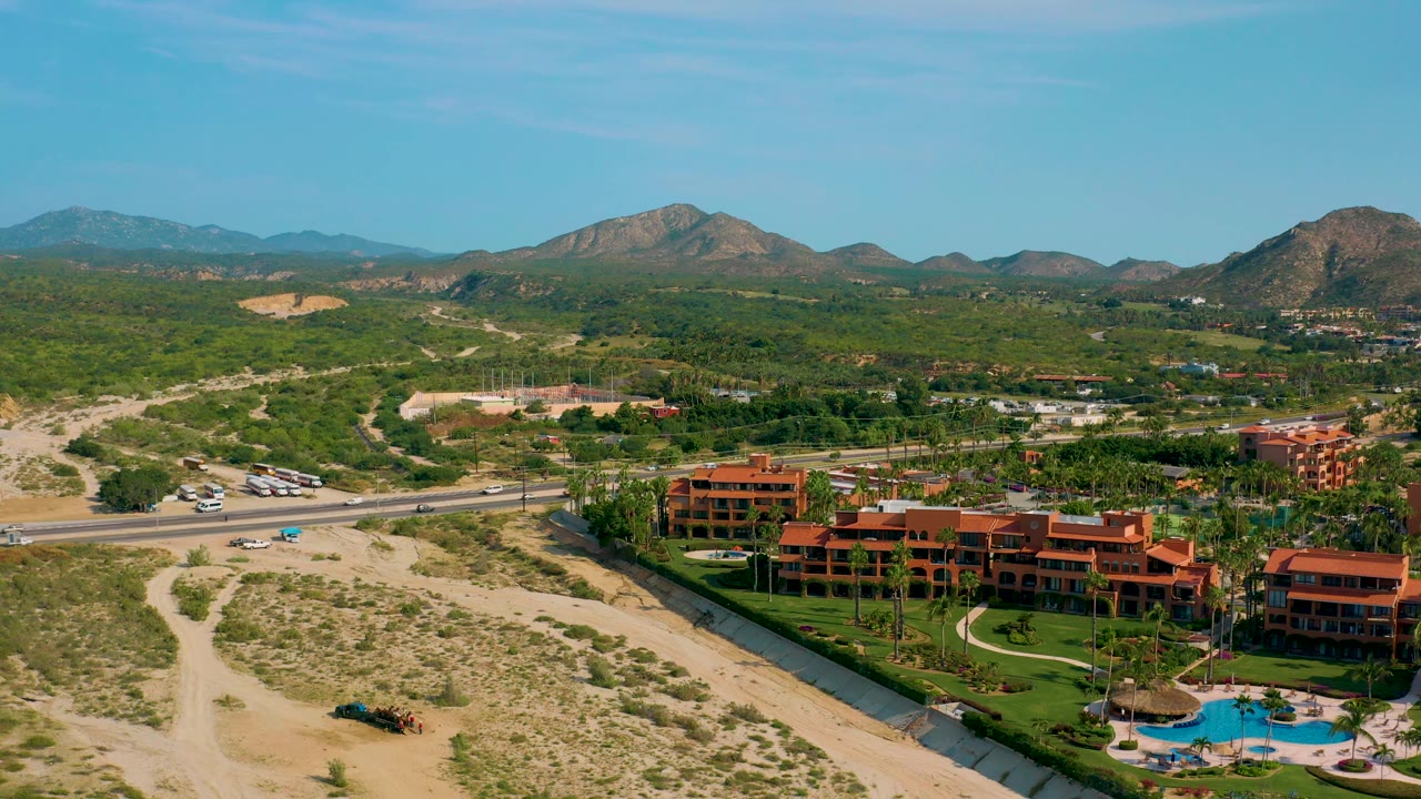 Drone frame over Baja California Sur resort architecture, with terracotta roofs and balconies near the bottom edge, a bright pool and palms, desert road cuts, highway traffic, green scrub hills, mountains, and no visible people.