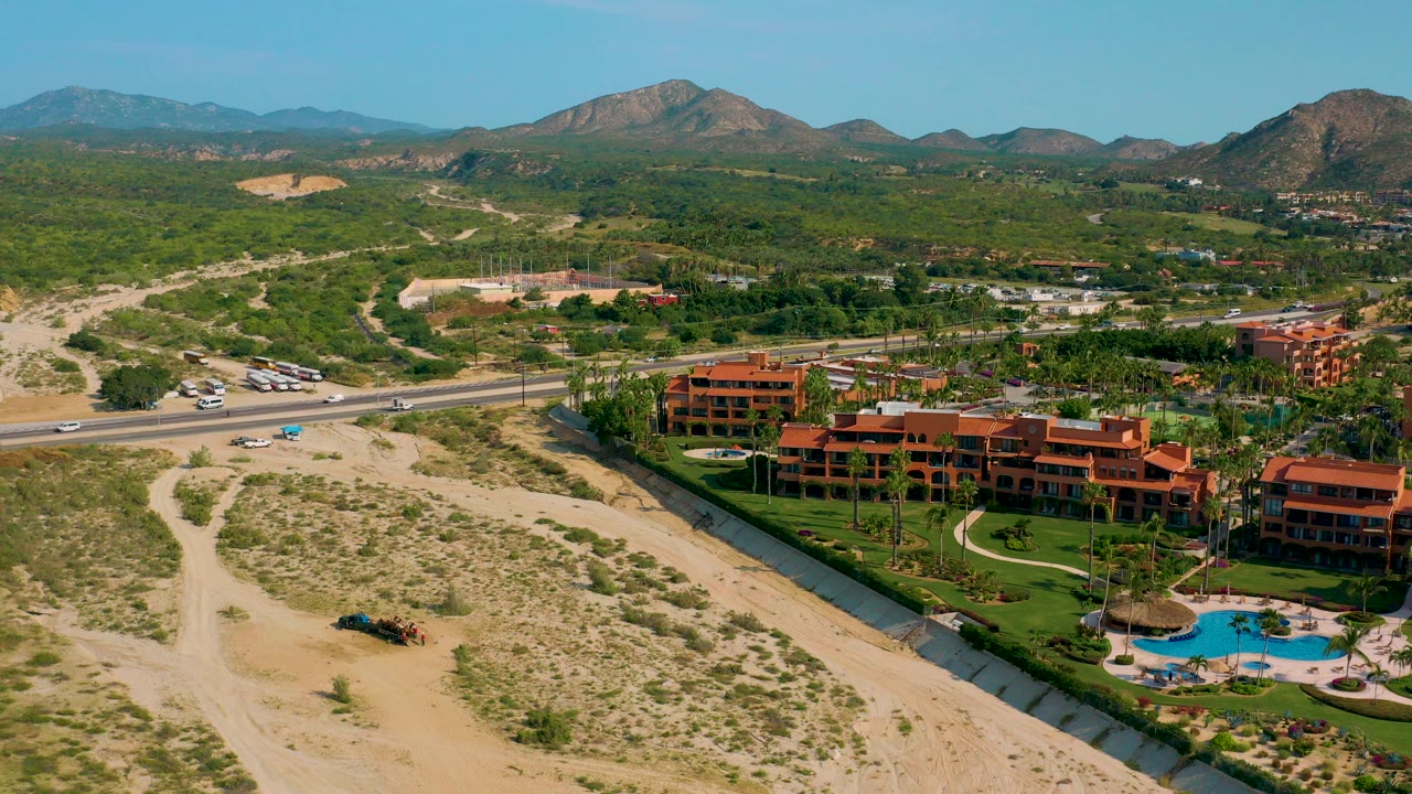 Wide aerial frame of Desert Park Natural Reserve in Baja California Sur, showing terracotta resort buildings, green grounds, sandy scrubland, a road, distant mountains, and no visible people.