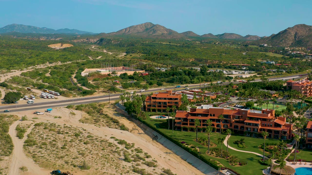 Aerial view in Baja California Sur with landscaped resort grounds in the lower frame, a parallel highway with small vehicles, desert scrub, dry washes, and distant mountains, with no visible people.