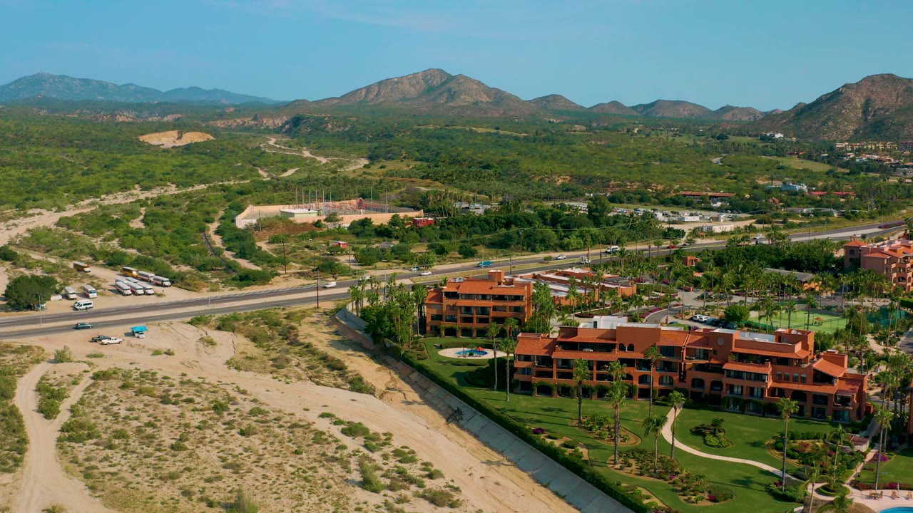 Baja California Sur aerial frame showing the resort shifted toward the lower right, a paved highway crossing the scene, sandy desert foreground, green vegetation, and rugged hills beyond, with no visible people.