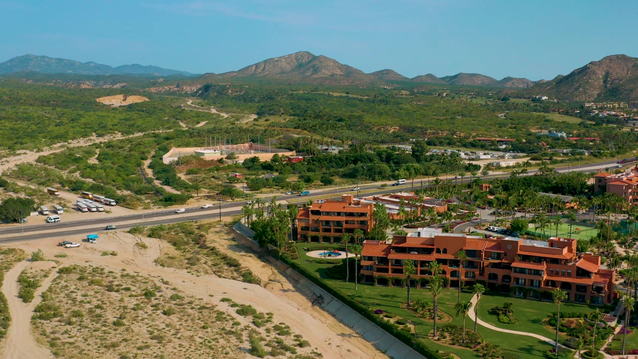 Wide aerial in Desert Park Natural Reserve with the highway running across the midground, resort buildings and lawns below it, open scrub desert behind it, and mountains across the horizon, with no visible people.
