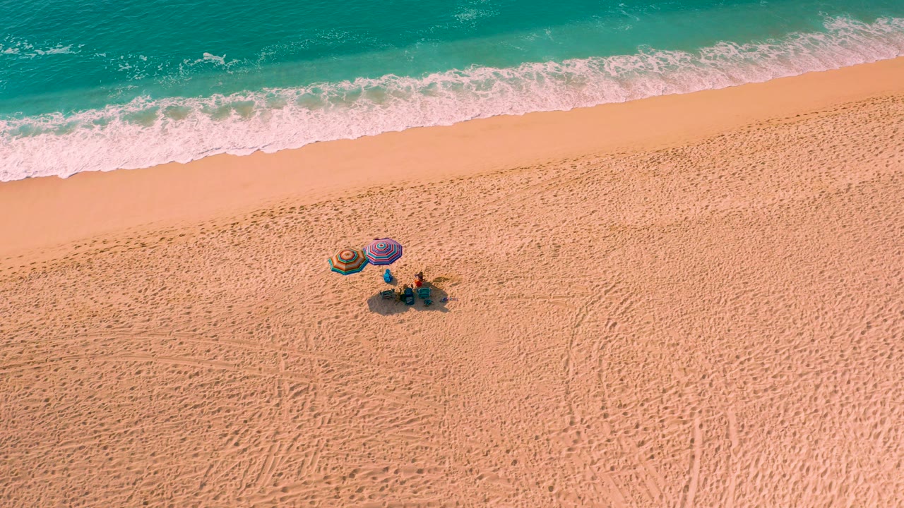 Overhead frame from Desert Park Natural Reserve, Baja California Sur, showing two small people beside colorful umbrellas on open sand below a turquoise wave line.