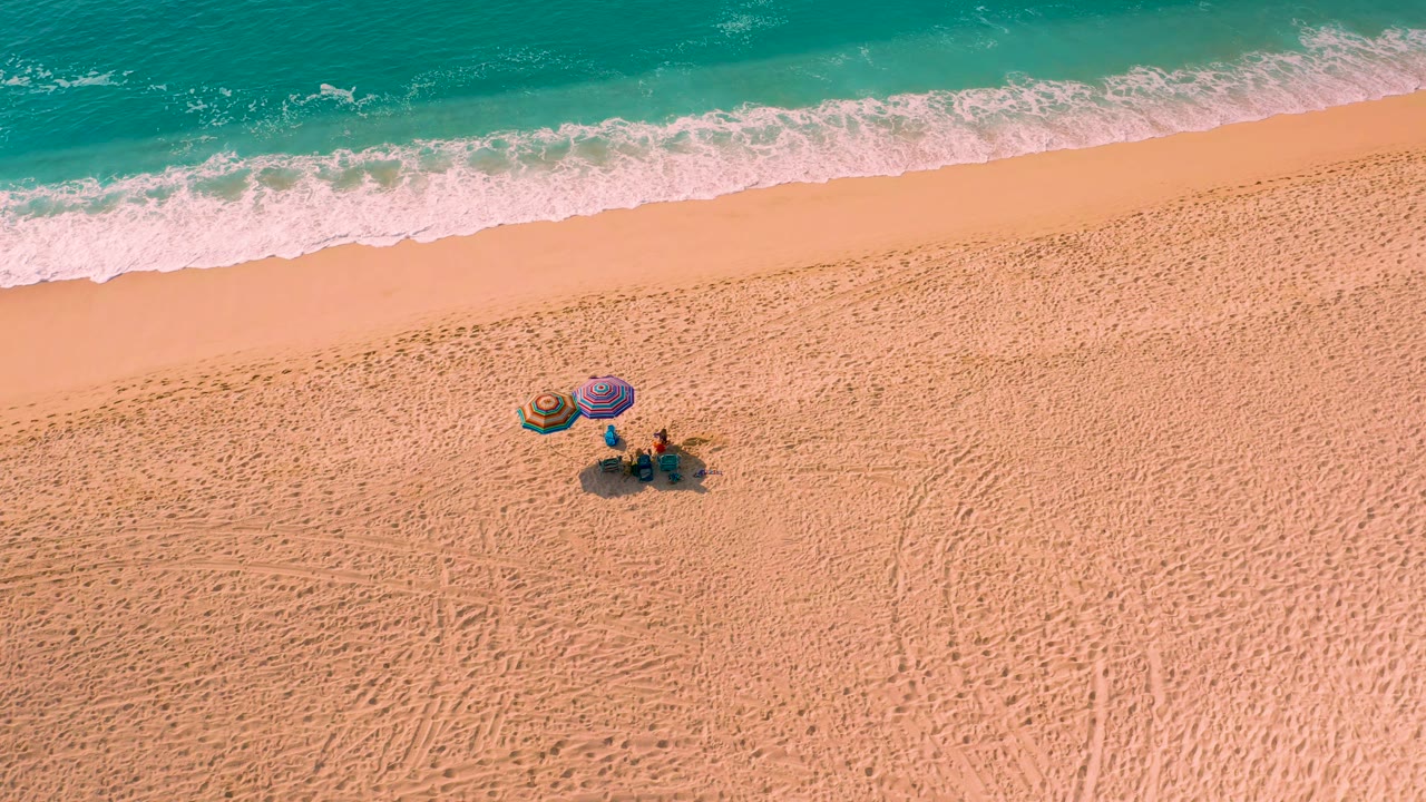 Aerial view of beach umbrellas and people on sand near rolling surf.