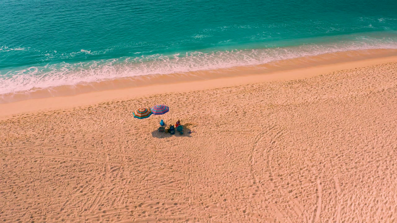 Aerial frame from Desert Park Natural Reserve in Baja California Sur showing a small group on open sand below two colorful umbrellas, with turquoise water and a thin surf line above them.