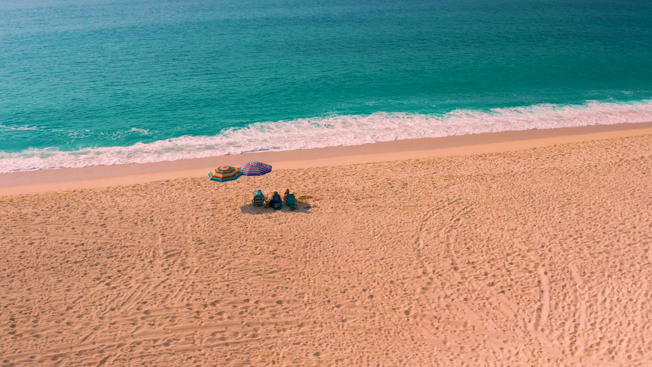 High-angle aerial view in Desert Park Natural Reserve, Baja California Sur, showing turquoise surf, golden sand, two colorful shade setups, and three seated people near the water.