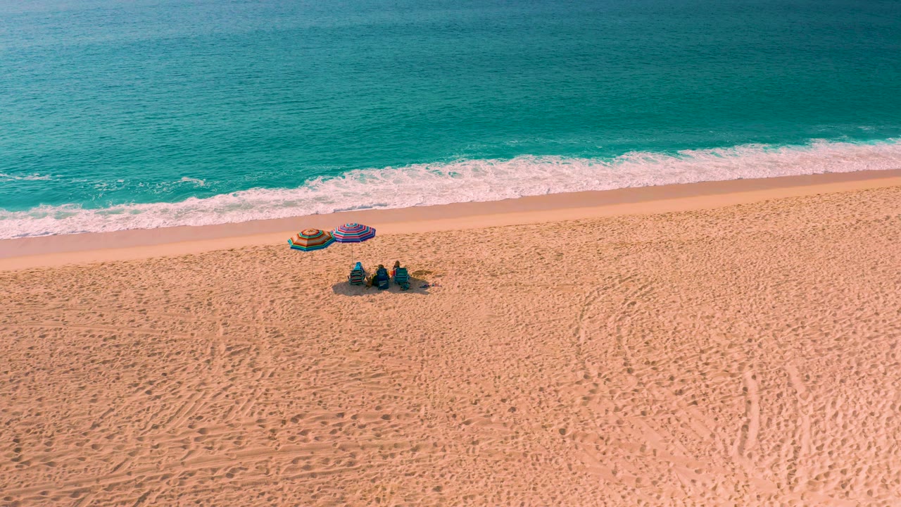Aerial shoreline with beach umbrellas and three people seated on sand.