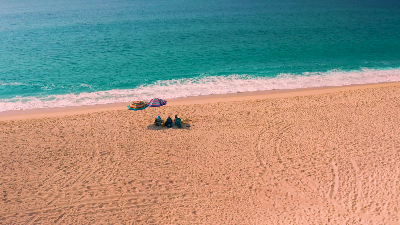 High aerial view over Desert Park Natural Reserve, Baja California Sur, where the surf band runs behind two small umbrellas and three seated beachgoers on textured sand.