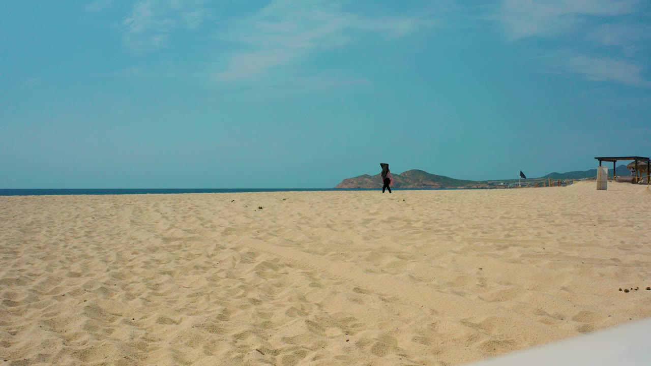 At Desert Park Natural Reserve in Baja California Sur, a lone person walks across a broad sandy beach with turquoise water, distant hills, and a small beach structure near the right edge.