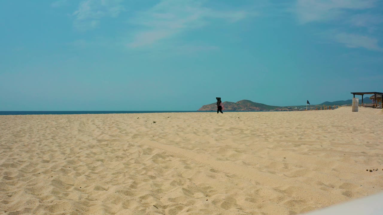 At Desert Park Natural Reserve in Baja California Sur, a single dark figure crosses sunlit sand while the blue ocean, low mountains, and right-side wooden shade structure remain in view.