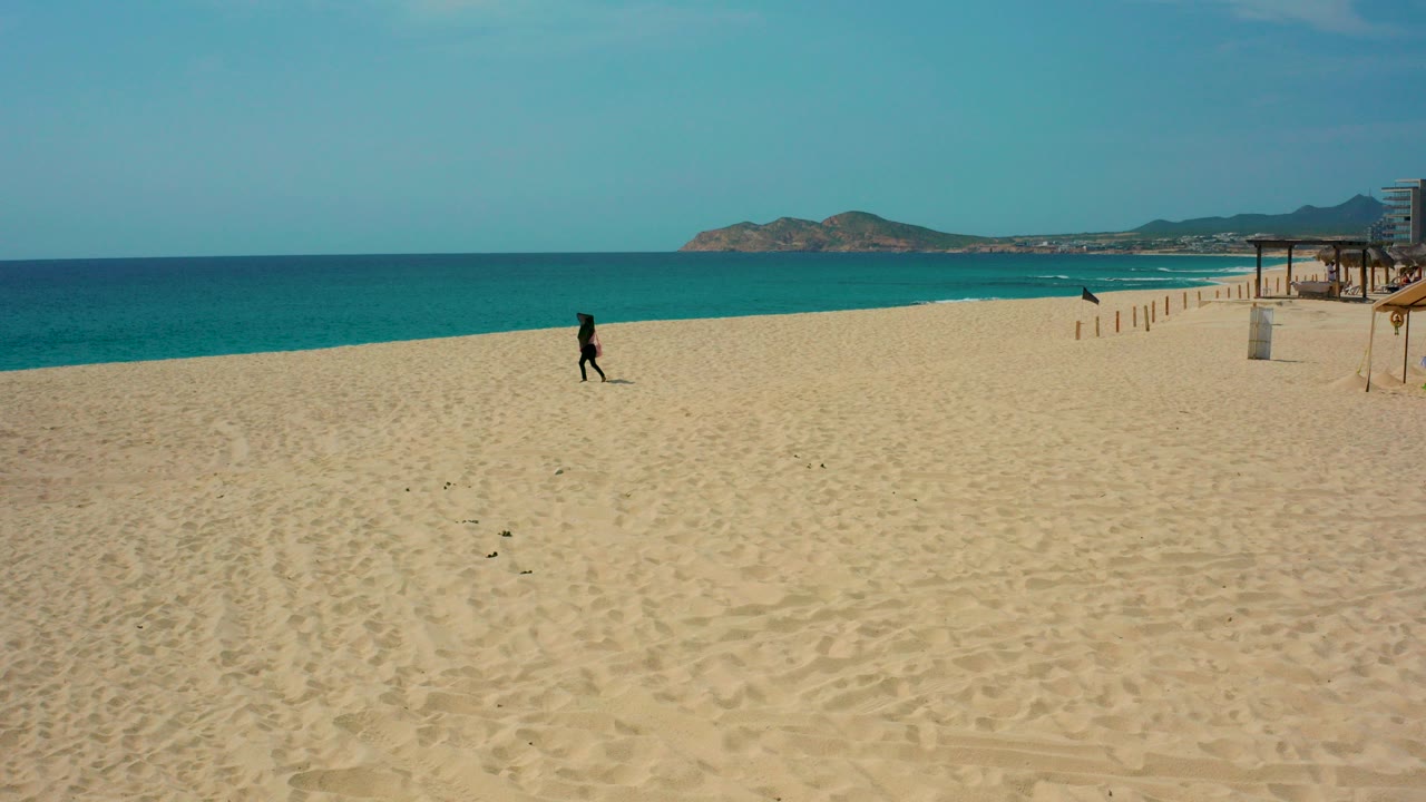 At Desert Park Natural Reserve in Baja California Sur, a lone runner moves across the open sand with turquoise ocean, distant landforms, and small beach structures visible.