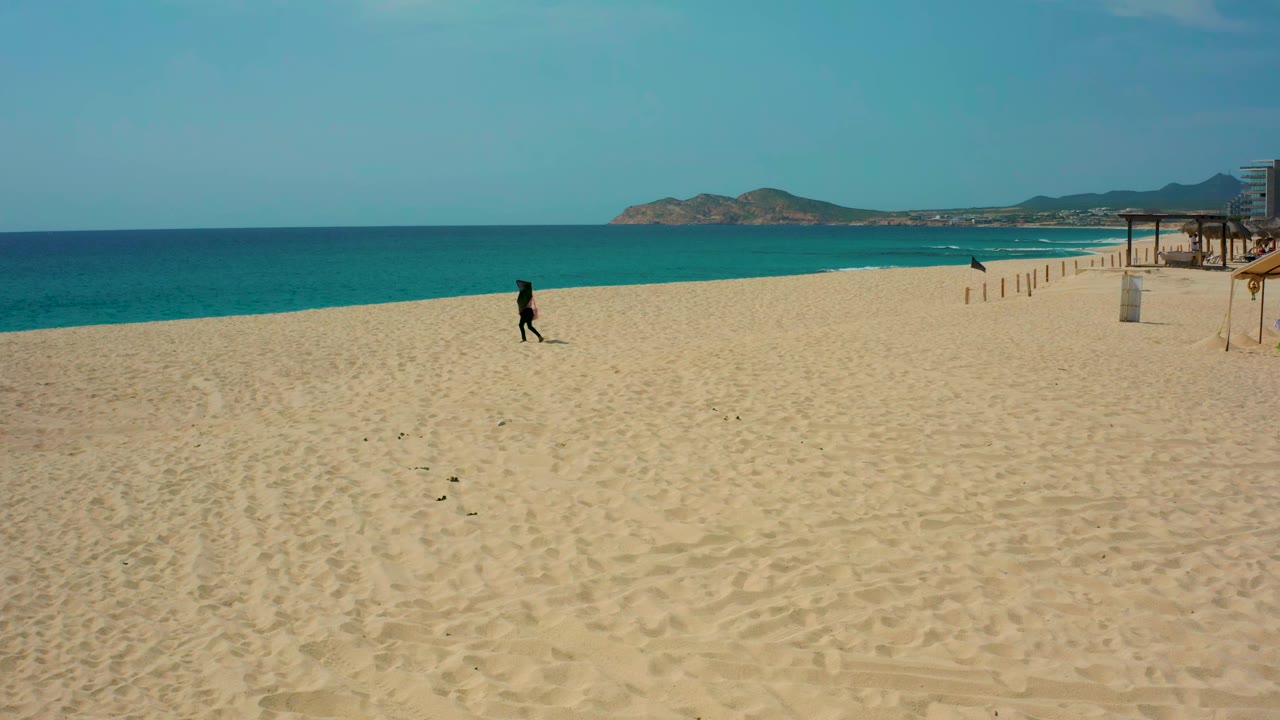 Runner crossing pale sand with ocean and distant hills behind.