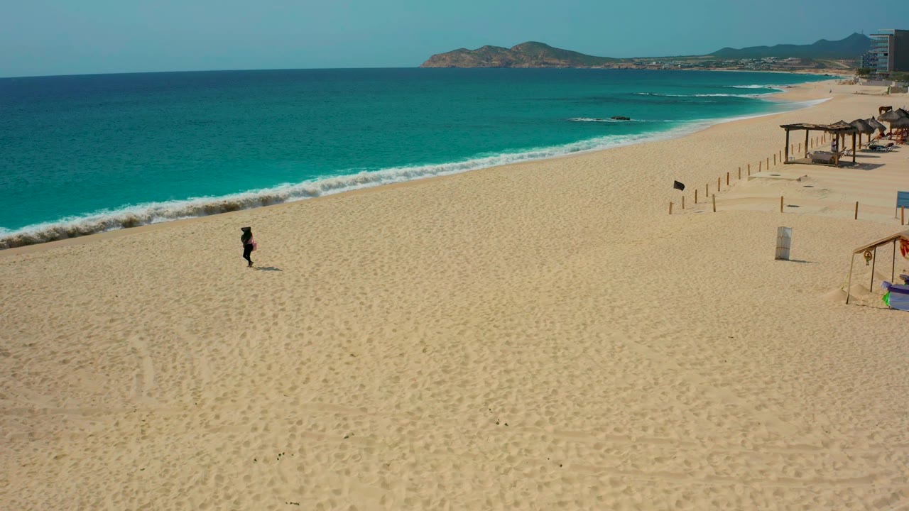 Aerial view of a lone walker on a broad sandy beach beside blue ocean.
