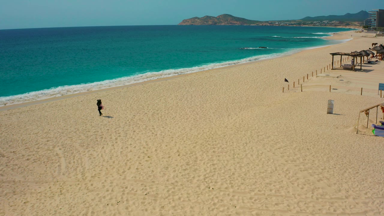 At 0:02, the person walks farther across the sandy foreground, with the surf curving behind them and small beach structures and posts along the right edge.