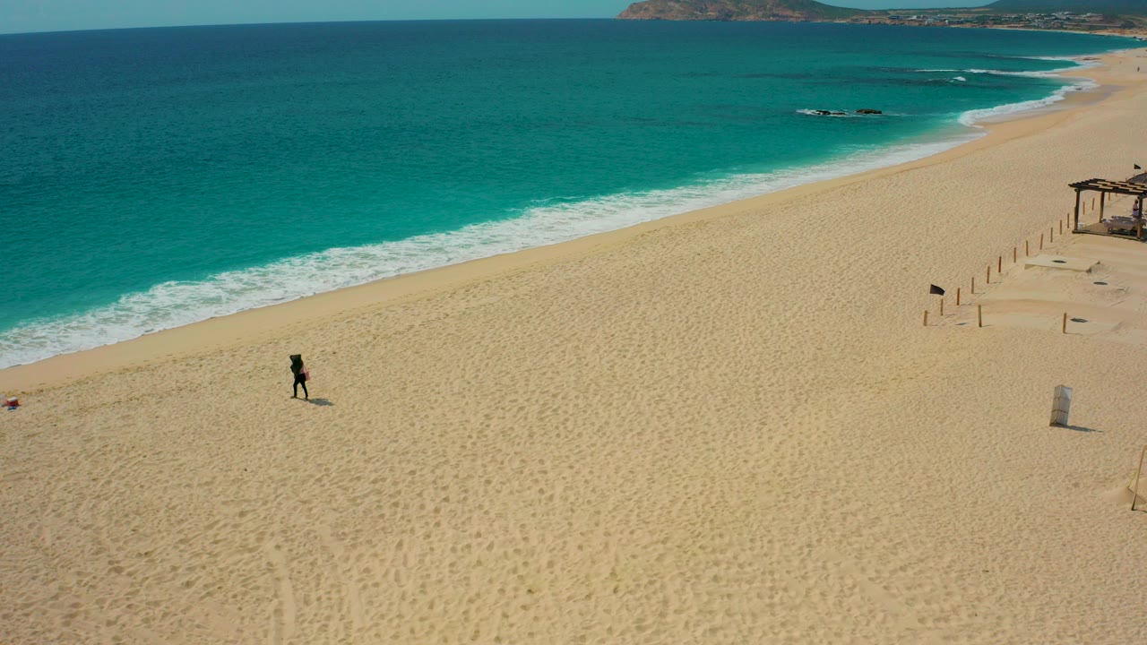 Aerial view in Desert Park Natural Reserve, Baja California Sur, showing one distant person on wide golden sand with turquoise ocean, white surf, posts, and a small wooden shade structure at the right edge.