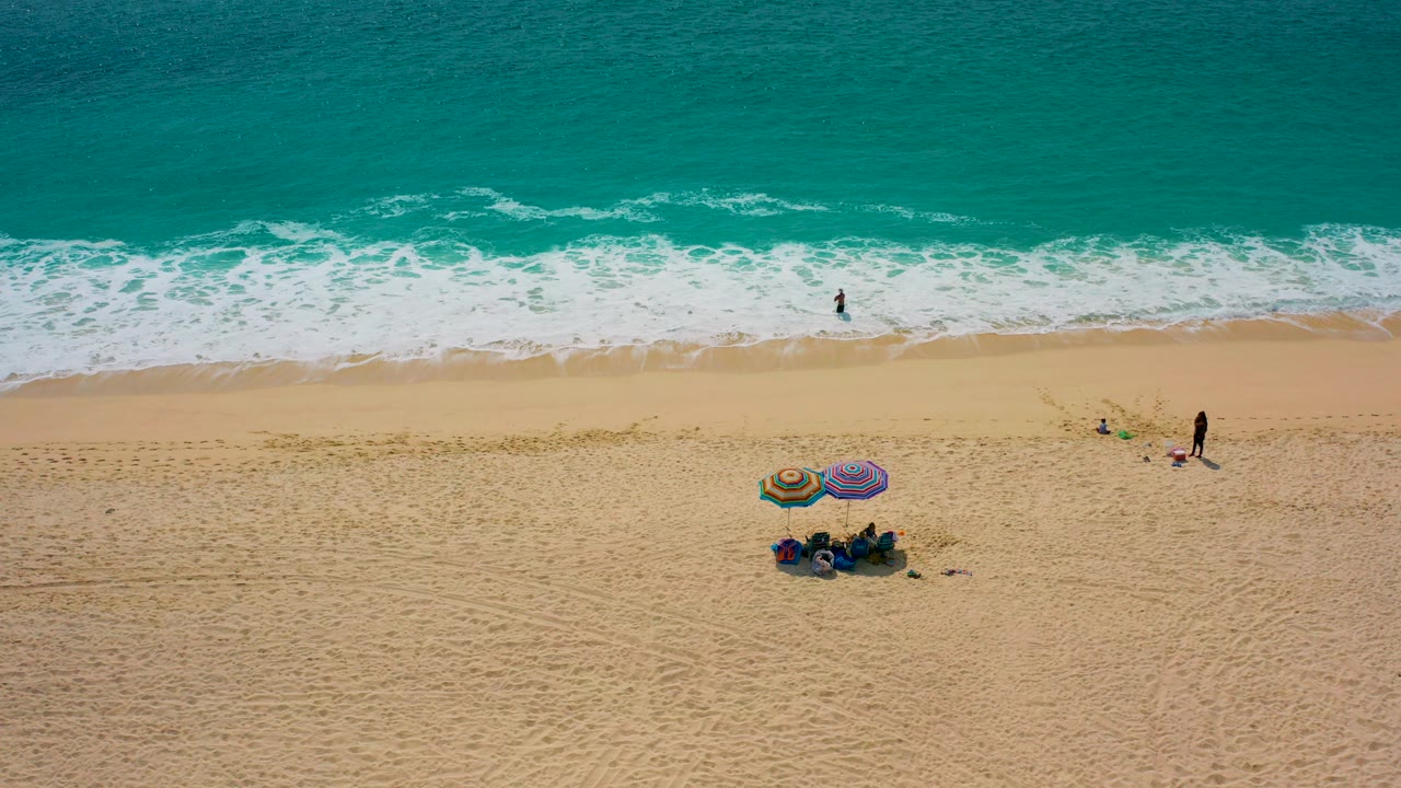 High-angle aerial frame from Desert Park Natural Reserve, Baja California Sur, Mexico, showing turquoise surf above golden sand, two colorful umbrellas, a small group below them, and distant people near the waterline.