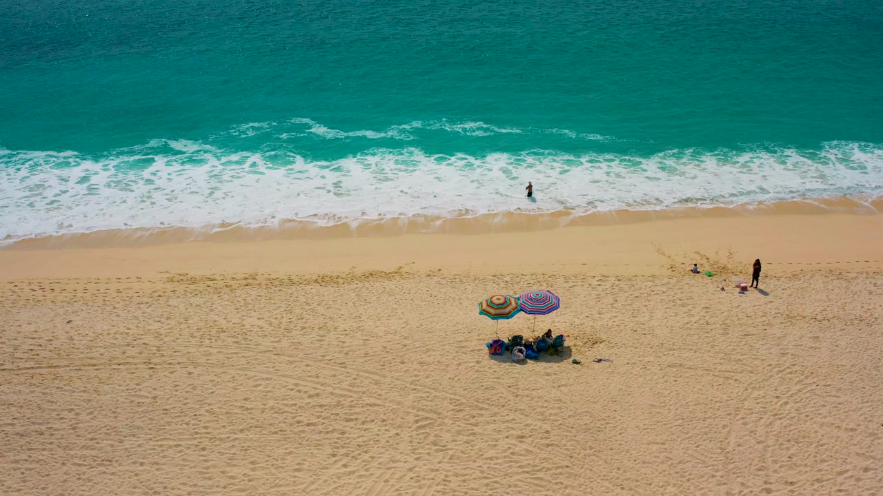 Drone view of waves, open sand, colorful umbrellas, and scattered beachgoers.
