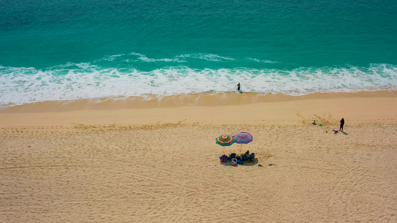 High aerial frame from Desert Park Natural Reserve, Baja California Sur, Mexico, showing a broad band of ocean, foamy waves, an open sandy beach, two umbrellas lower in frame, and several small human figures.