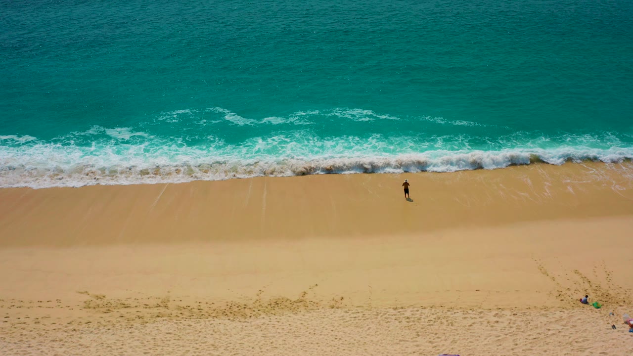 Aerial view in Desert Park Natural Reserve, Baja California Sur, showing a lone surfer walking on golden sand below turquoise water and a thin band of breaking foam.