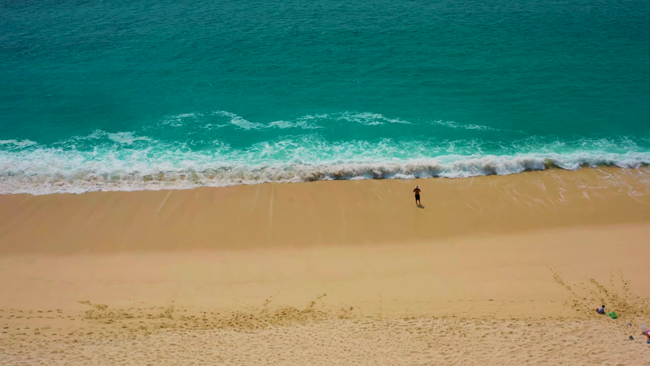 Lone surfer walks parallel to the surf on a sandy beach.