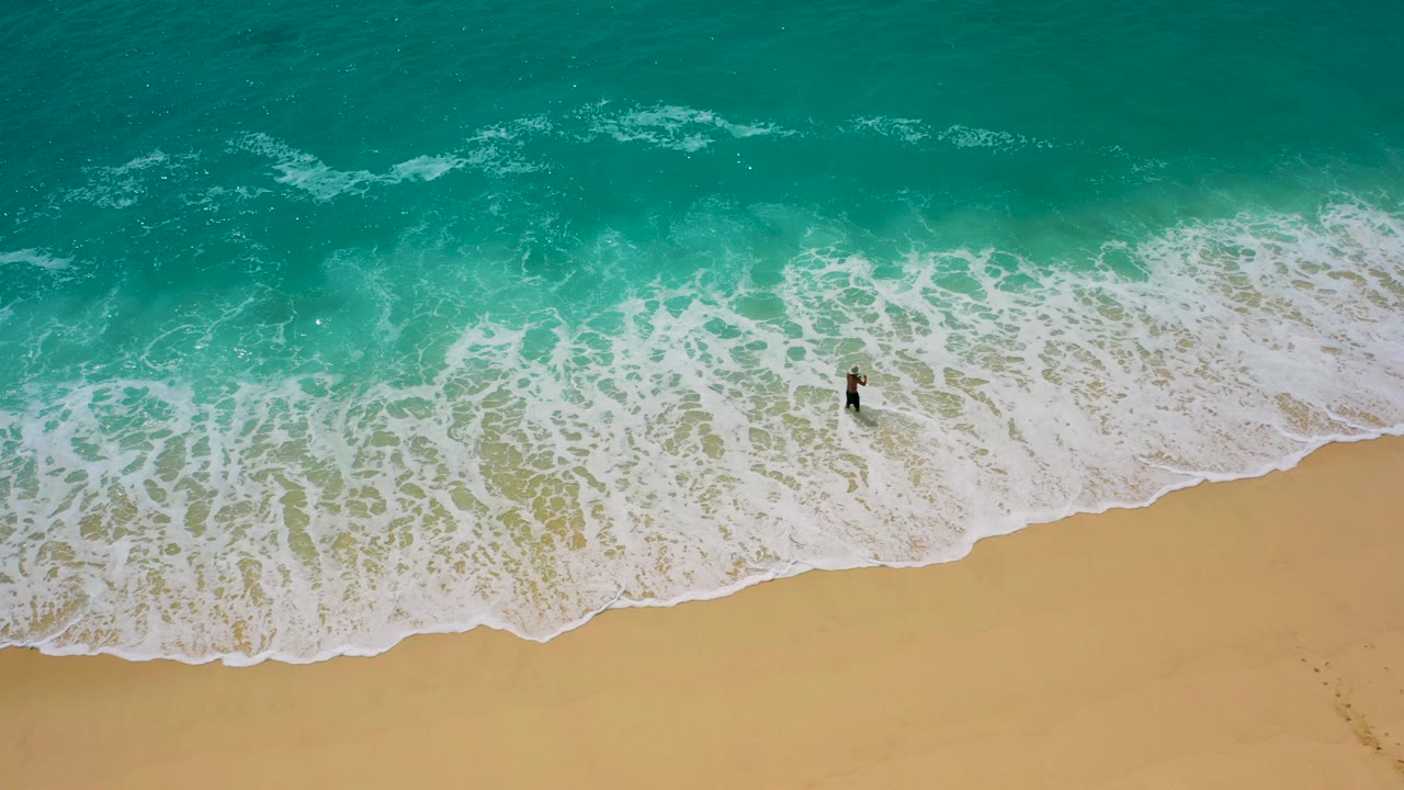 Top-down aerial frame from Baja California Sur, Mexico, showing a lone surfer standing in foamy shorebreak between turquoise ocean and golden sand.