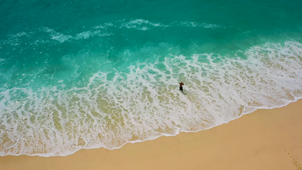 Aerial top-down frame from Baja California Sur, Mexico, showing a lone surfer near the upper edge of the sandy beach as white water spreads diagonally across the shoreline.