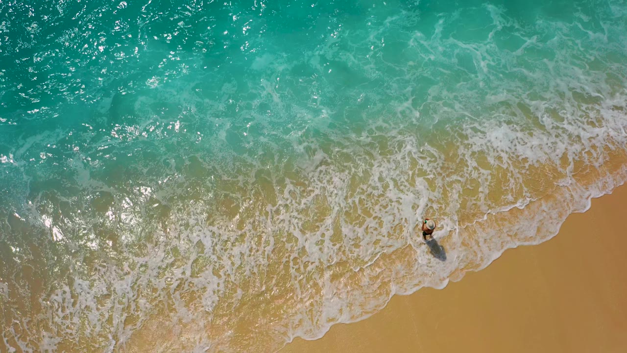 Top-down frame from Desert Park Natural Reserve, Baja California Sur, where the surfer stands near a wider lace of foam spreading over the tan beach.