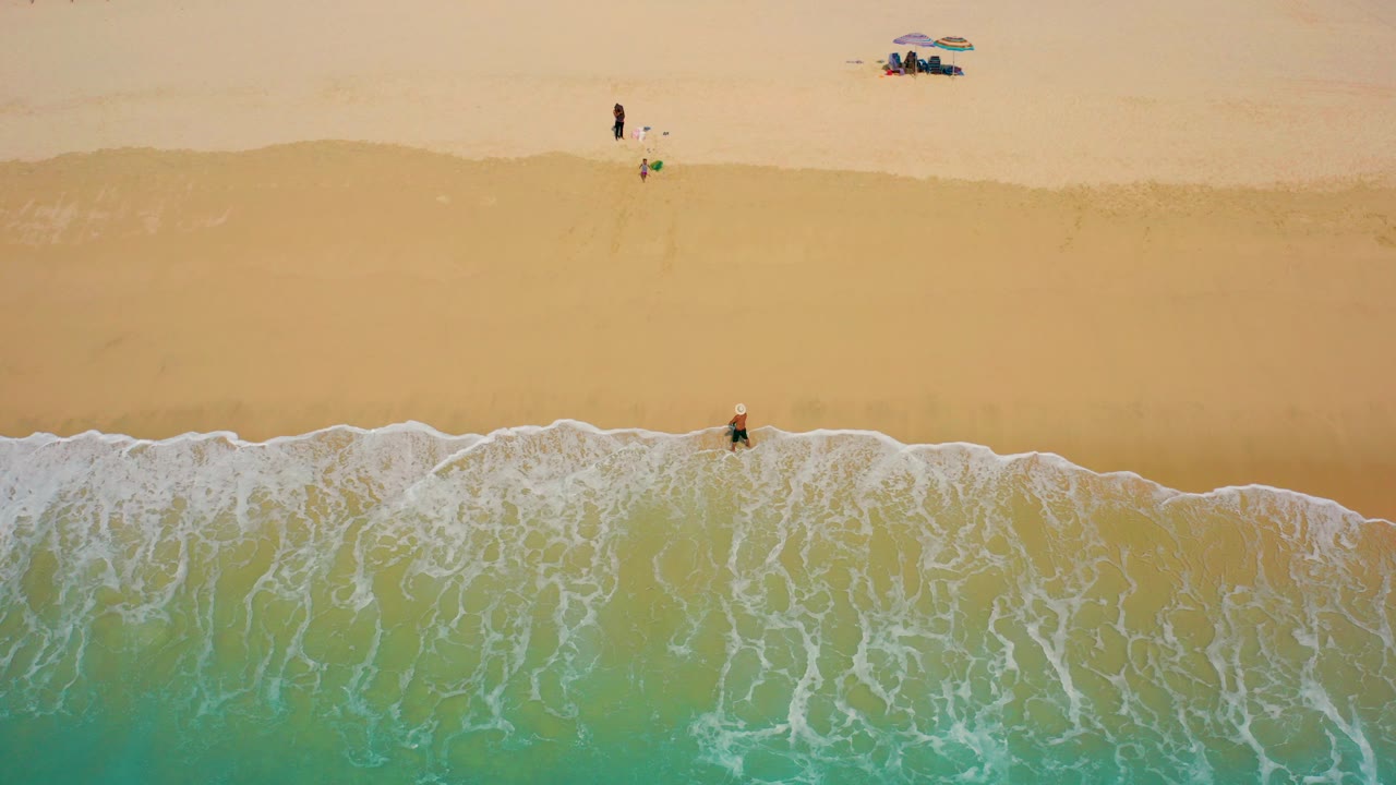 Overhead view in Desert Park Natural Reserve, Baja California Sur, with a lone surfer at the foam line, turquoise water below, and small beachgoers near an umbrella on the upper sand.