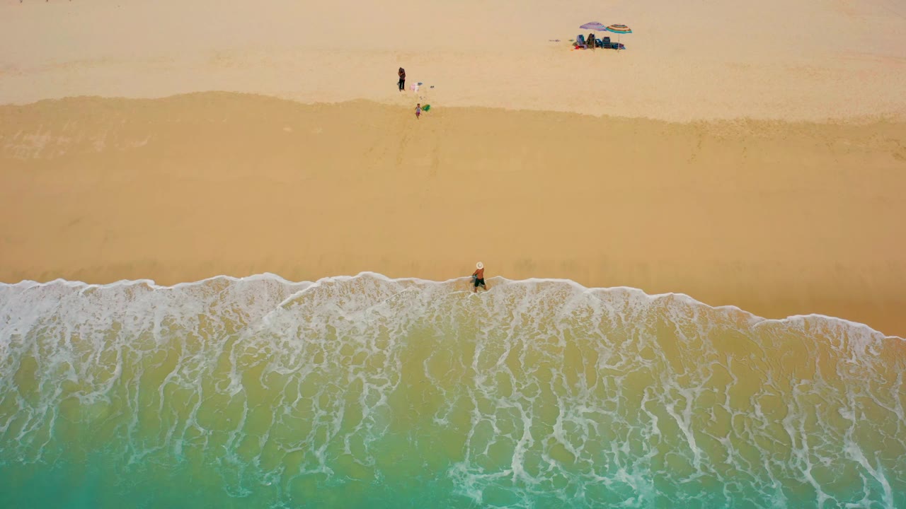 Top-down Baja California Sur shoreline frame with the surfer still near the breaking wave, thin white foam spreading across the sand, and a small umbrella group near the top of frame.