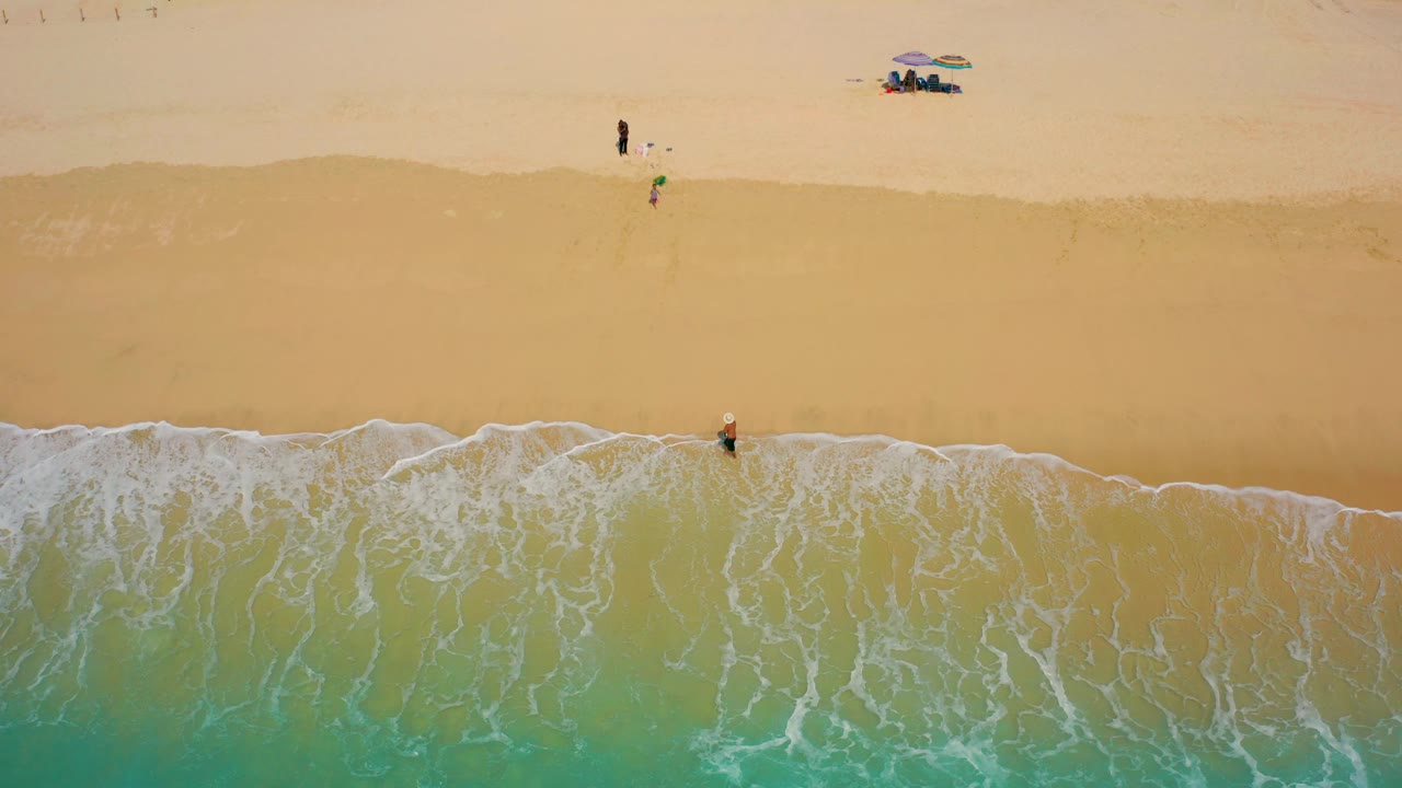 Aerial view at Desert Park Natural Reserve showing thicker white surf patterns across the lower frame, the lone surfer near the waterline, and distant beachgoers on the pale sand above.
