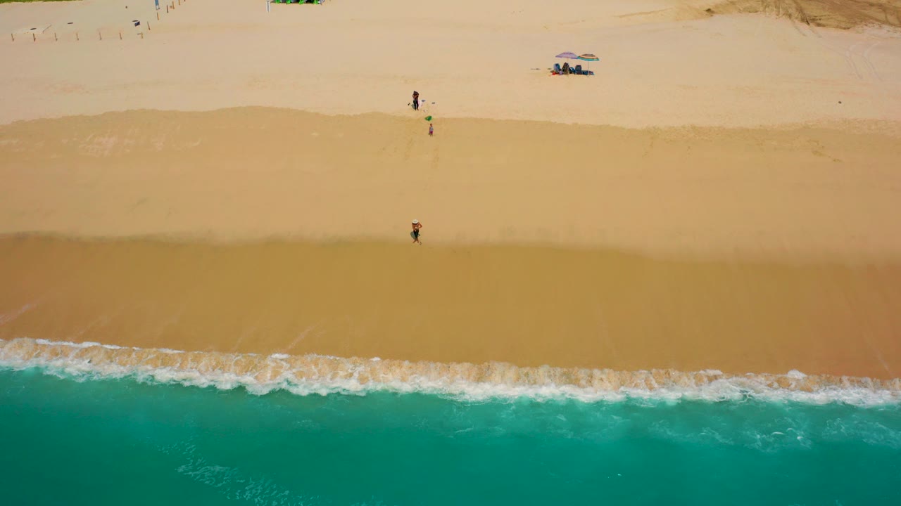 Overhead aerial frame from Desert Park Natural Reserve in Baja California Sur, Mexico, showing teal ocean below, white surf on tan sand, tiny beach walkers, umbrellas, and vehicles near the dunes.