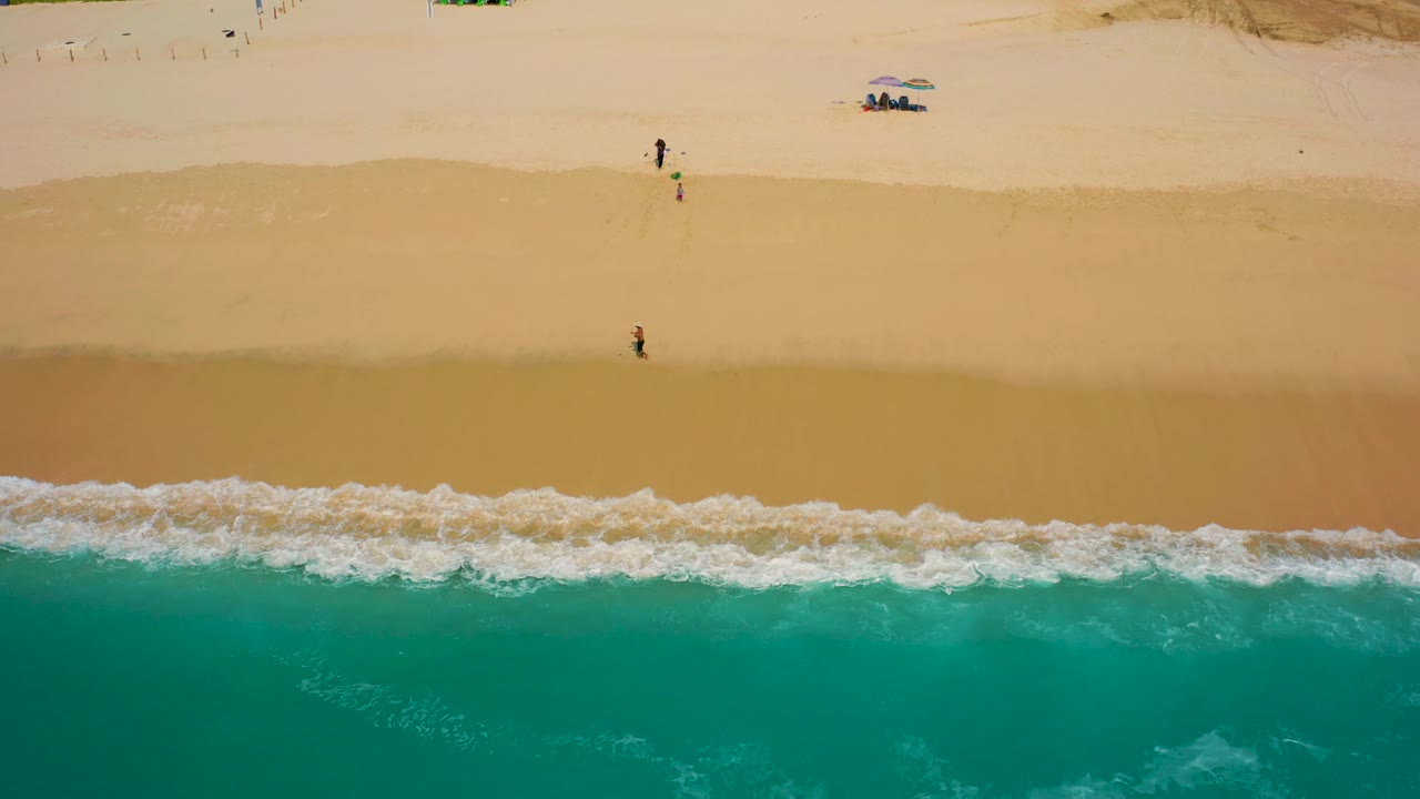 Drone-style overhead frame in Baja California Sur showing foamy surf across the lower edge, open sand through the center, two small beachgoers, umbrellas, and vehicles near the top of the beach.