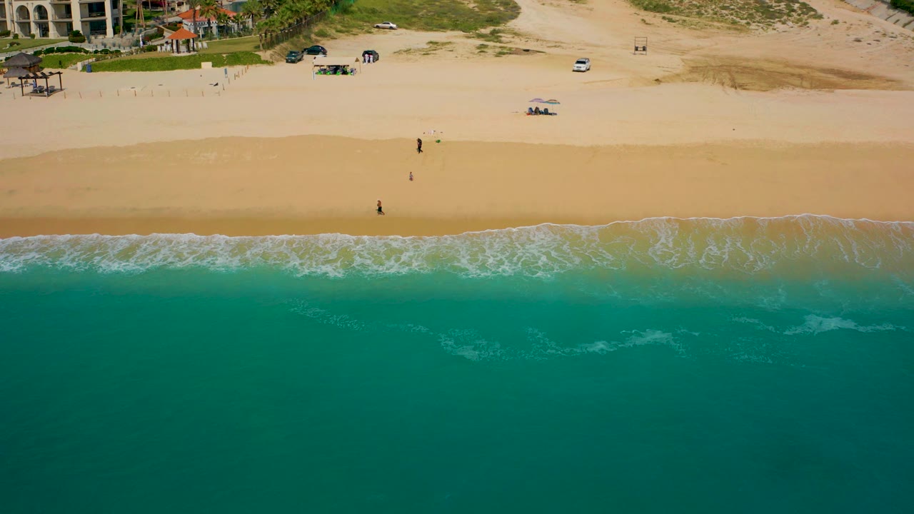 High-angle aerial view in Baja California Sur, Mexico, with teal ocean filling the lower frame, a narrow surf line, a broad sandy beach, and resort-style buildings at the top.