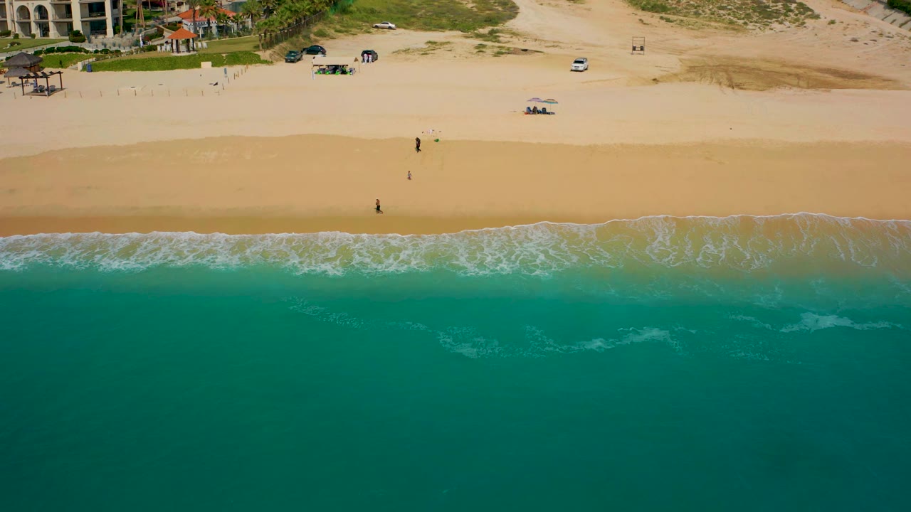 Aerial frame over Baja California Sur coastline showing more open turquoise water below, a thin white wave band, pale sand, sparse beach objects, and resort buildings near vegetation.