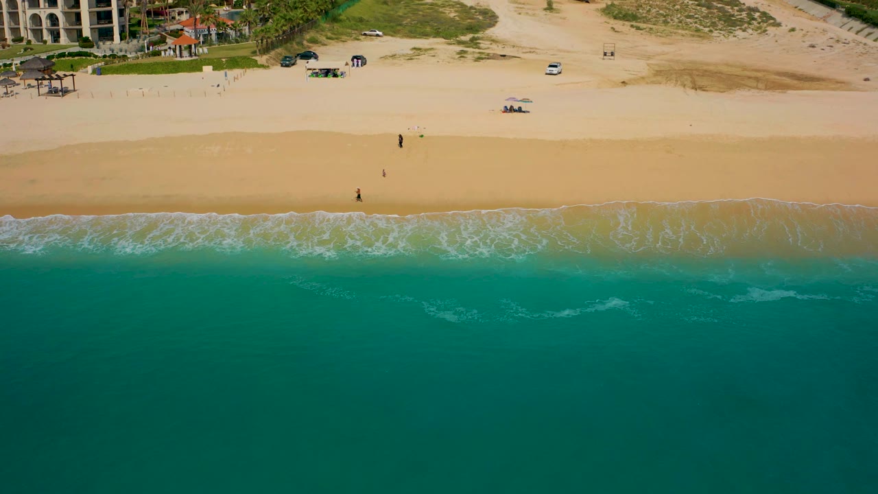 Baja California Sur aerial frame with the surf line running horizontally across the image, open beach above it, patches of vegetation, and resort-style buildings near the upper corners.