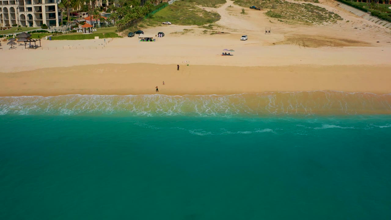 Aerial beach view with darker teal water, pale sand, and resort buildings along the upper edge.