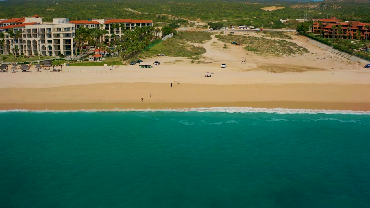 Aerial view in Baja California Sur showing a long pale beach between turquoise water and resort blocks, with arid green hills and low mountains behind the buildings.
