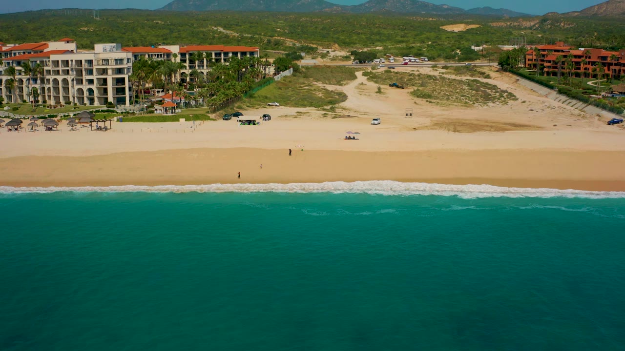 High aerial frame of Desert Park Natural Reserve coastline, with ocean filling the lower half, a thin surf edge, a broad beach, resort buildings, and scrub-covered hills in the distance.