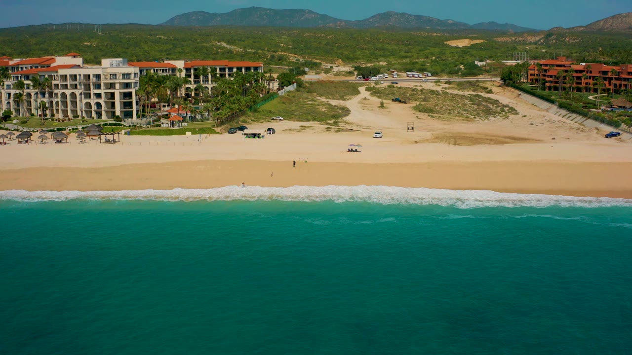 Baja California Sur aerial frame with low surf reaching the sand, a resort complex set back from the beach, sandy access areas, and mountains across the upper background.