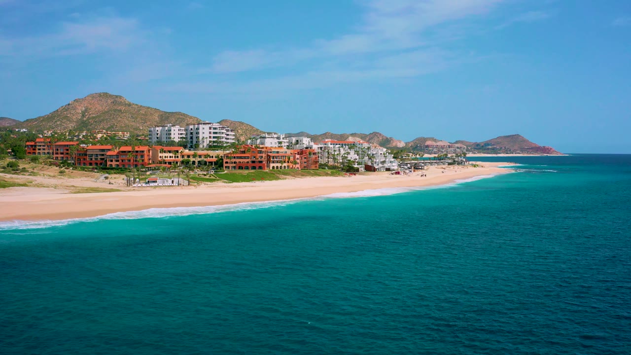 High-angle aerial view over Desert Park Natural Reserve coastline, with blue-green ocean taking most of the frame, a thin surf edge, beachfront hotels, hills, and no visible people.