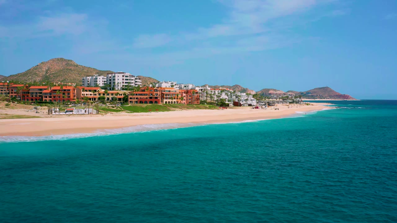 Wide aerial view across clear blue ocean to the sand and multi-story beachfront hotels in Baja California Sur, Mexico, with no visible people.